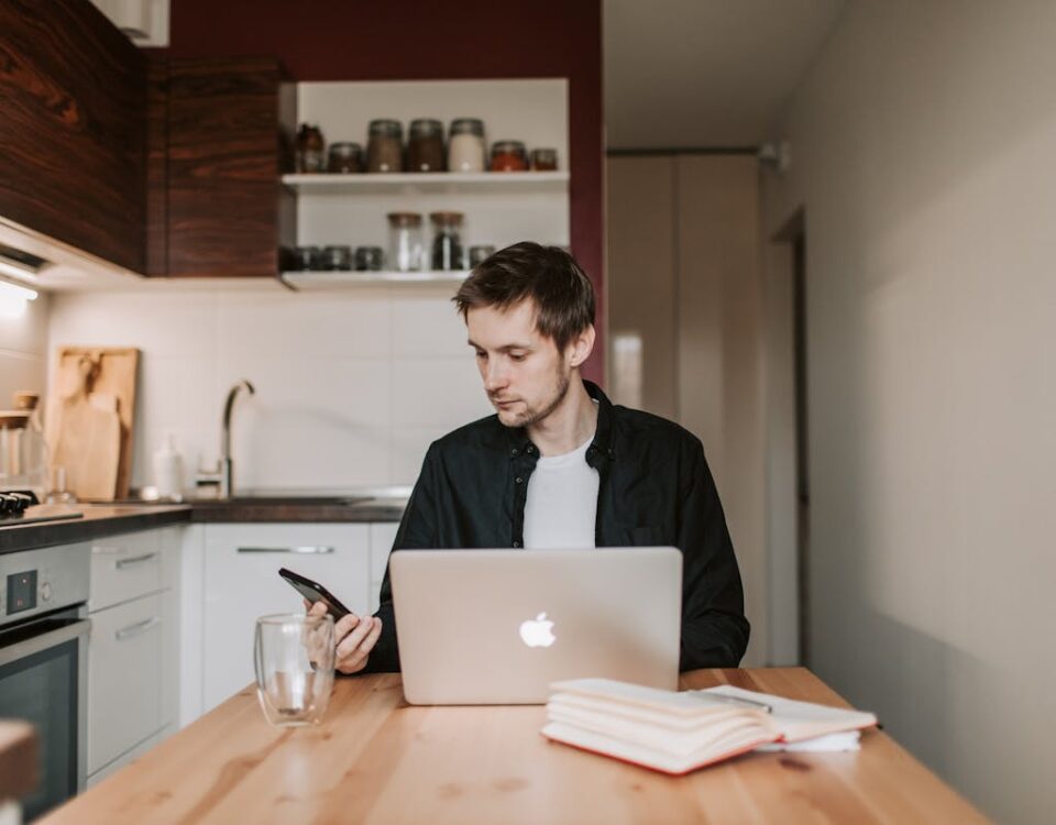 Como o WhatsApp Business pode aumentar suas vendas? Young male remote employee in casual wear chatting on cellphone while sitting at wooden table in front of open netbook and copybook with glass of water in flat