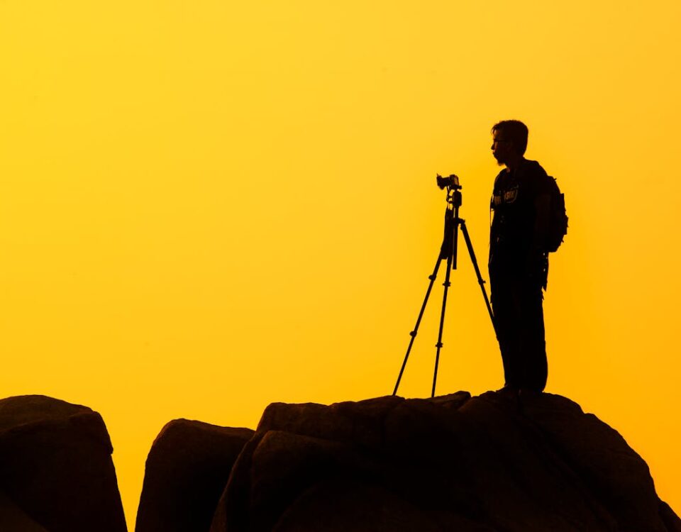 Estratégias para artistas monetizarem sua arte online Silhouette of a lone photographer with tripod on a rocky cliff against a golden sunset sky.