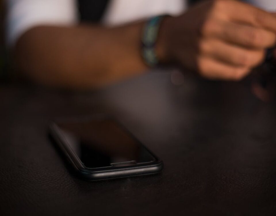 Como não cair em falso Golpe do PIX errado? A smartphone rests on a table with a person holding an object in the blurred background, in a dark indoor setting.