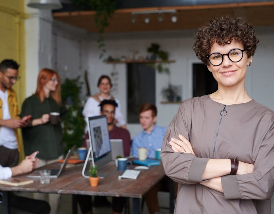 A gestão que reduz perdas e aumenta o lucro Smiling businesswoman with curly hair stands confidently in a modern office space with colleagues.