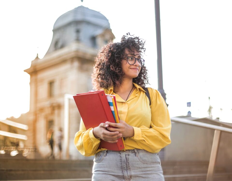 Como criar um método de estudo que cabe no orçamento Smiling female student with curly hair and glasses holding books on campus steps.