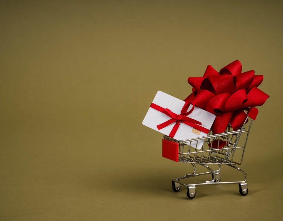 Planejamento de Marketing de Fim de Ano para Pequenas Empresas Mini shopping cart containing a gift card adorned with a red bow on a neutral background, ideal for retail concepts.