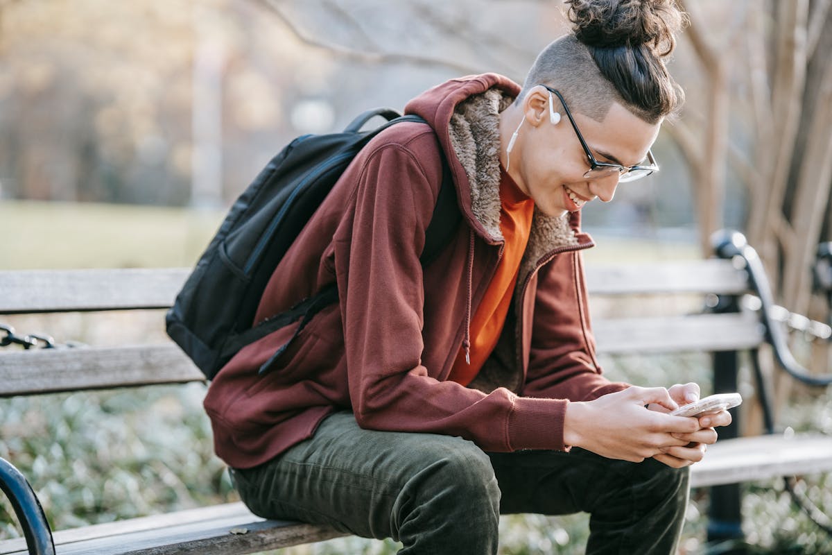 A young man using his smartphone while sitting on a park bench on a sunny autumn day.