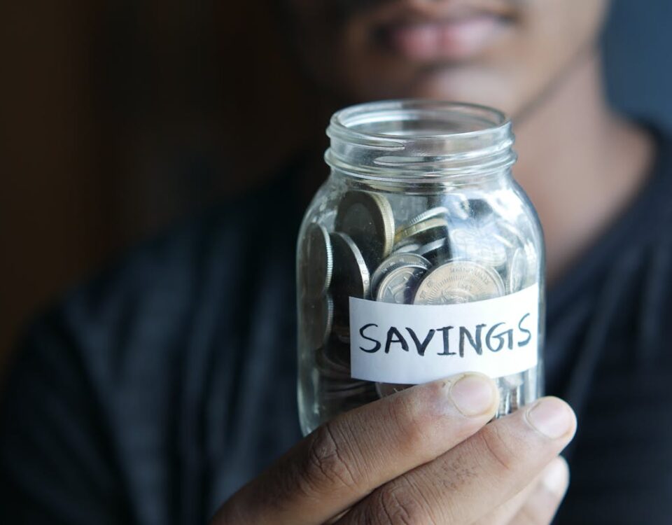 Como criar um fundo de emergência sem sacrificar o lazer A close-up image of a person's hand holding a jar full of coins labeled 'Savings'.