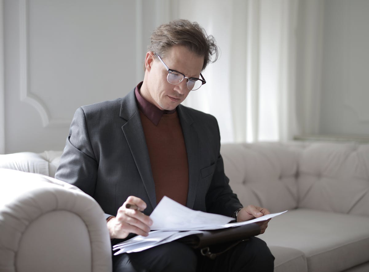 Seguro de vida: Custo ou investimento financeiro? Elegant adult man in jacket and glasses looking through documents while sitting on white sofa in luxury room