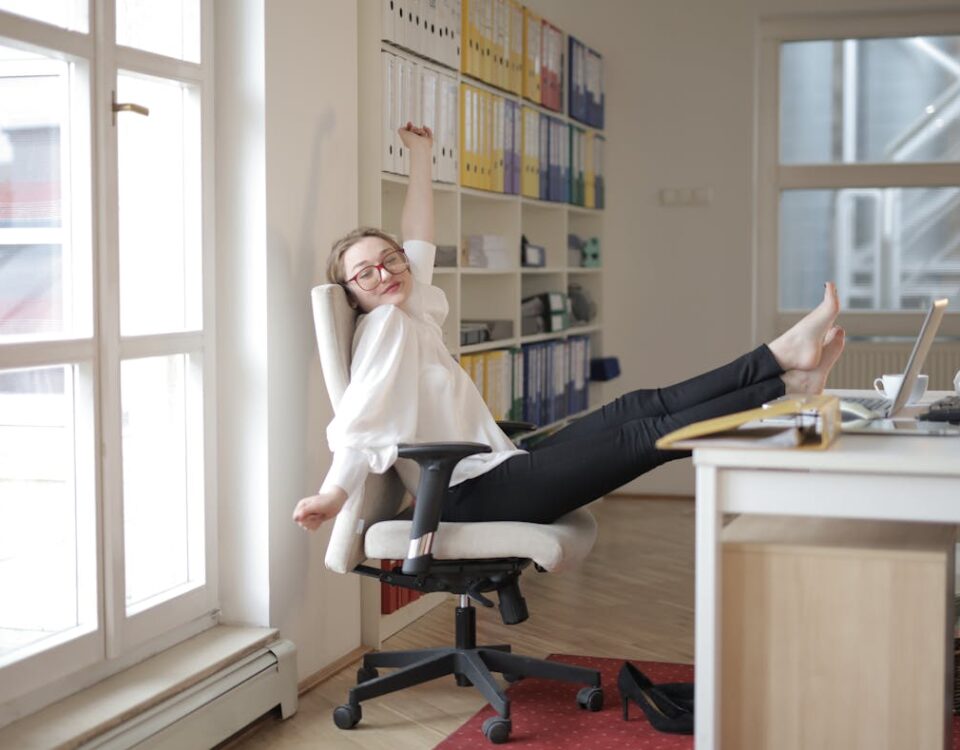É possível economizar investindo em saúde no trabalho? Cheerful woman relaxing in a modern office, barefoot with feet on the desk, enjoying a tranquil break.