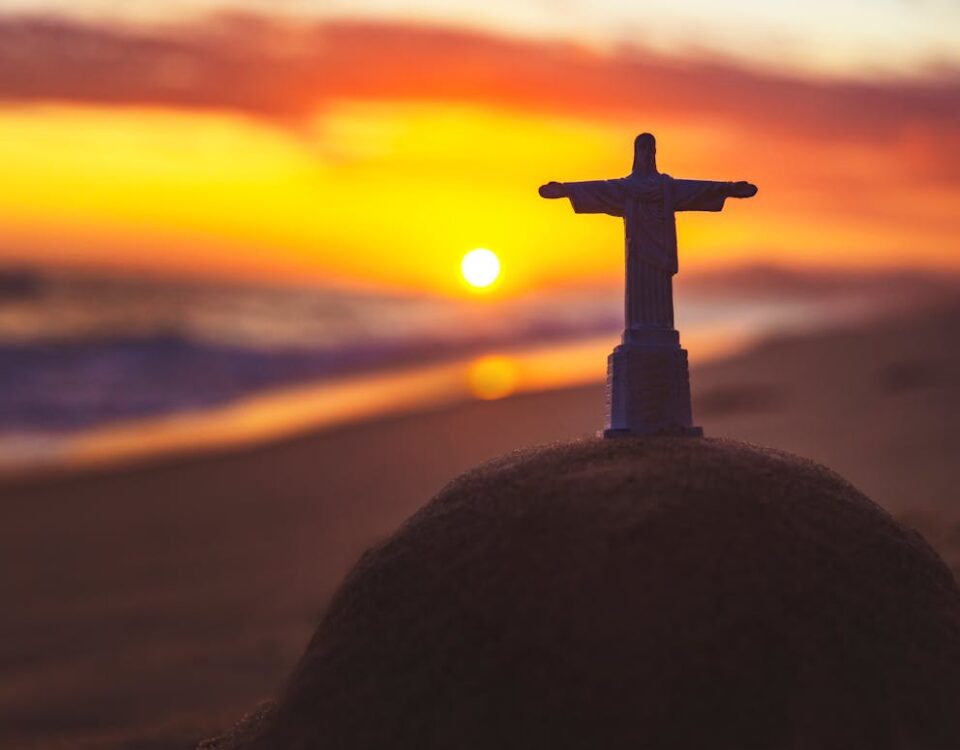 Como economizar na compra de lembranças de viagem A miniature Christ the Redeemer figurine on sand during a vibrant beach sunset, evoking spirituality.