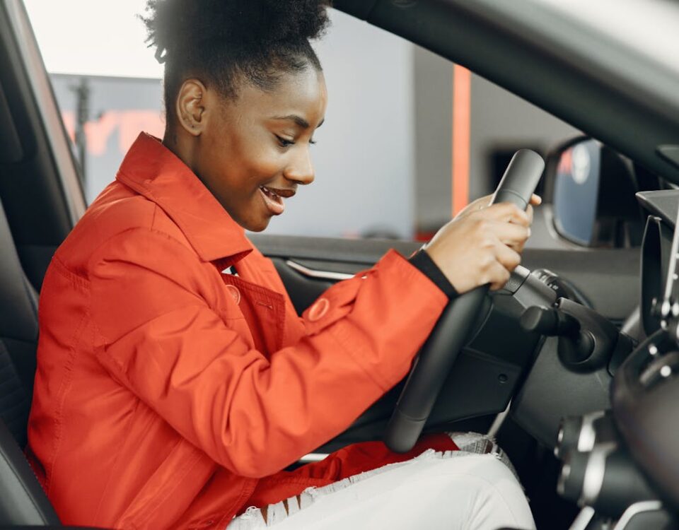 Quanto custa tirar a CNH em 2025? Young woman in an orange jacket enthusiastically trying out a car's steering wheel.