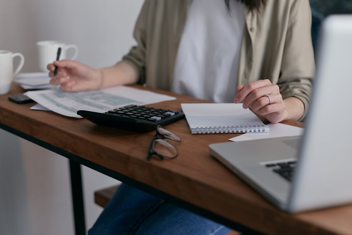 Automação comercial: Como reduzir custos e aumentar lucros? A woman manages finances at home, using a laptop and calculator on a wooden desk.
