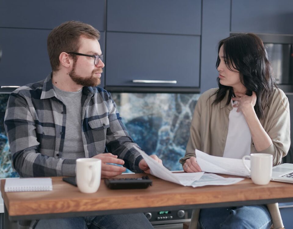 Como o despachante ajuda a economizar com taxas A young couple sitting at a table discussing bills and financial plans in a modern kitchen.
