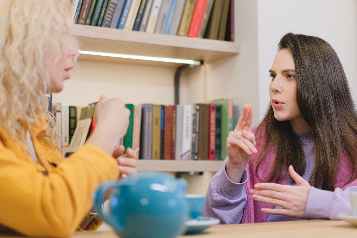 Vale a pena investir em cursos de idiomas? Side view of female friends sitting at table in university library and using sign language