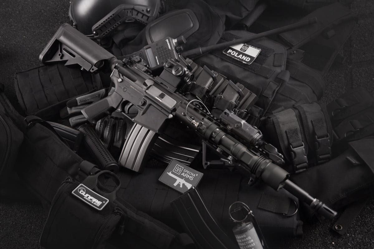 Close-up of tactical military gear including a rifle, radio, and vest on a dark background.