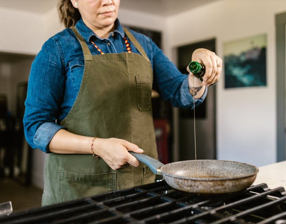 Cozinhar em casa: Quanto você economiza realmente? A home chef in an apron pours olive oil into a frying pan on a stove.