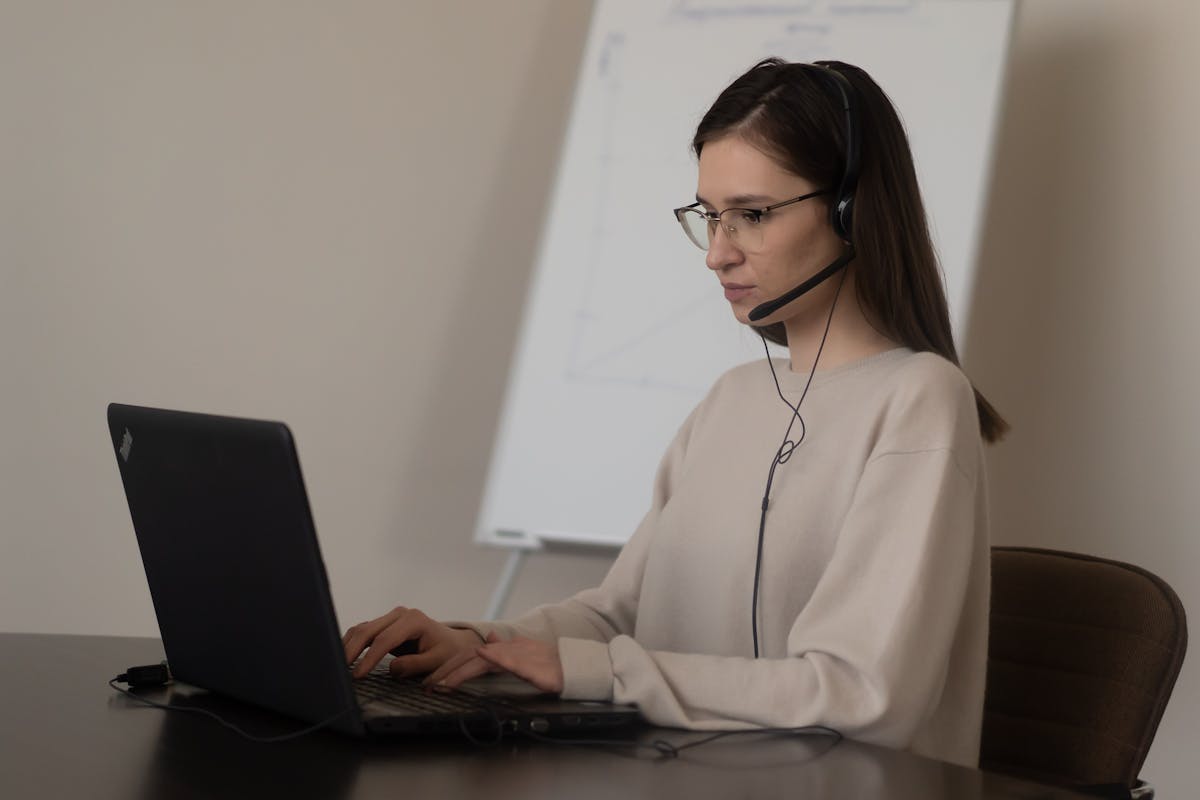 Atendimento eficiente pode ser mais barato do que você imagina Serious young woman in casual outfit with eyeglasses sitting on chair at table and working on laptop with headphones with microphone near whiteboard in bright workplace