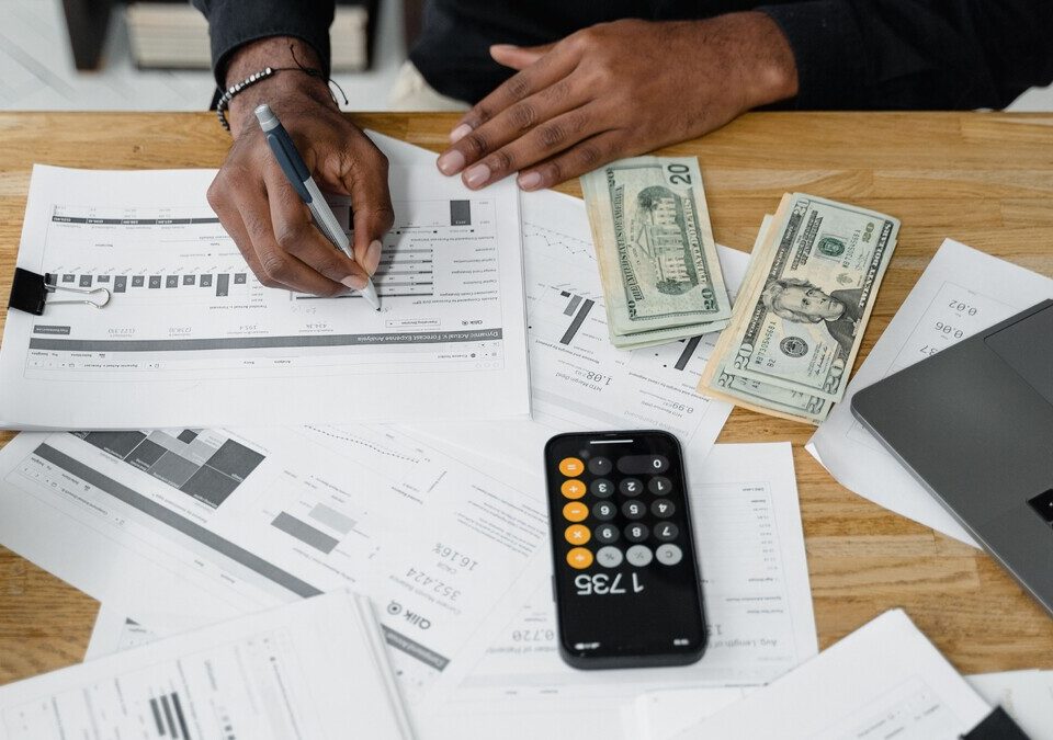 Pessoa analisando relatórios financeiros, calculadora e dinheiro sobre uma mesa.