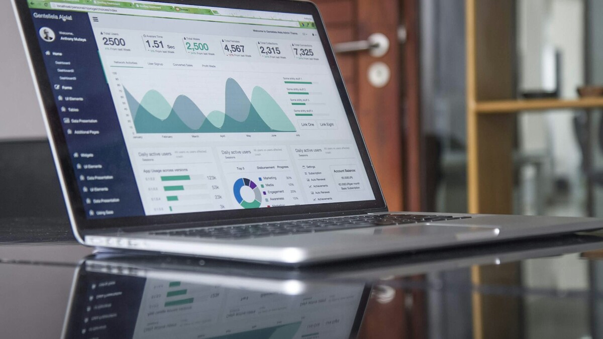 Notebook aberto sobre mesa de vidro exibindo um painel com gráficos de área e indicadores